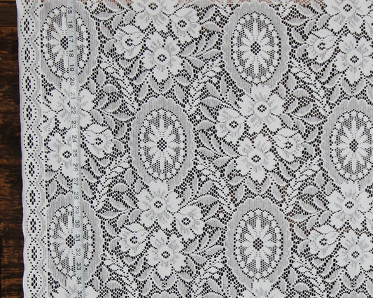 White floral lace fabric with intricate embroidered patterns and a scalloped border, laid flat on a dark wooden surface.