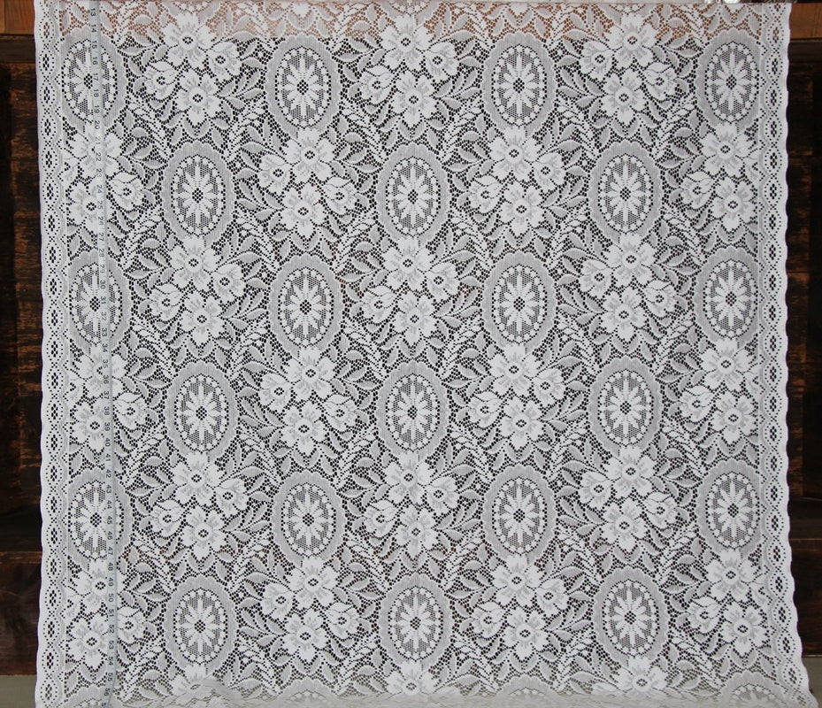 White floral lace textile with repeating oval medallion motifs and scalloped edges, laid flat against a dark wood background.