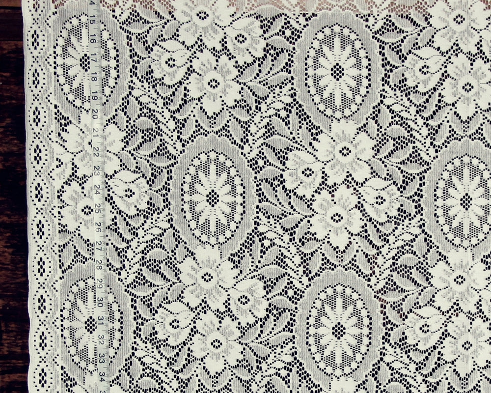 White floral lace fabric with intricate embroidered patterns and a scalloped border, shown laid flat against a dark wood background.