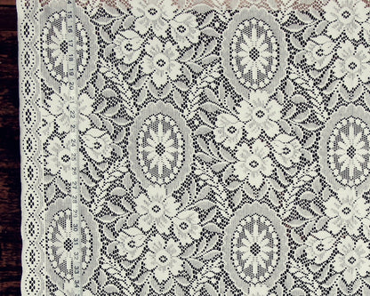 White floral lace fabric with intricate embroidered patterns and a scalloped border, shown laid flat against a dark wood background.