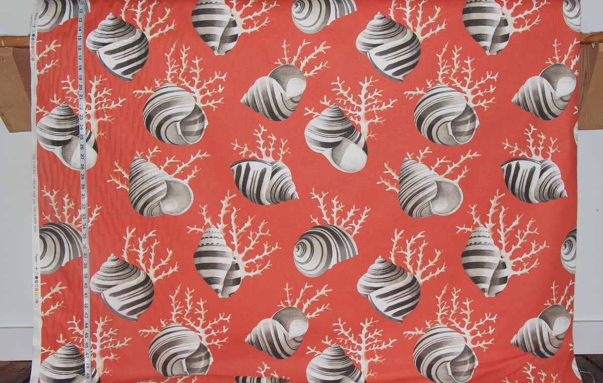 A close-up of a printed fabric with a coral and seashell pattern in orange-red, gray, and white, with a measuring tape visible along the left edge.