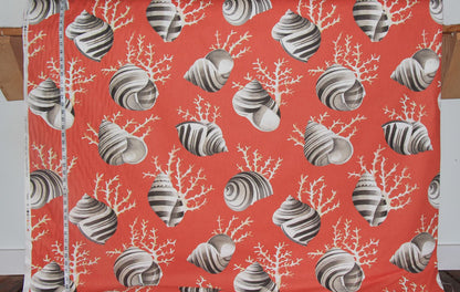 A close-up of a printed fabric with a coral and seashell pattern in orange-red, gray, and white, with a measuring tape visible along the left edge.