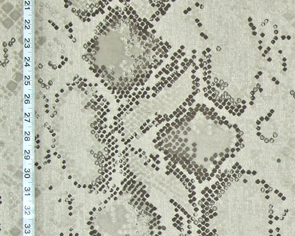 A close-up of a snakeskin-patterned fabric with a light beige base and dark brown to black dotted scales, featuring a white ruler on the left side for scale.