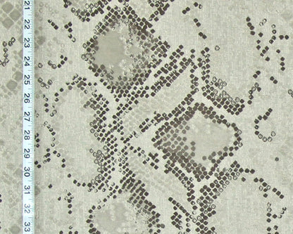 A close-up of a snakeskin-patterned fabric with a light beige base and dark brown to black dotted scales, featuring a white ruler on the left side for scale.