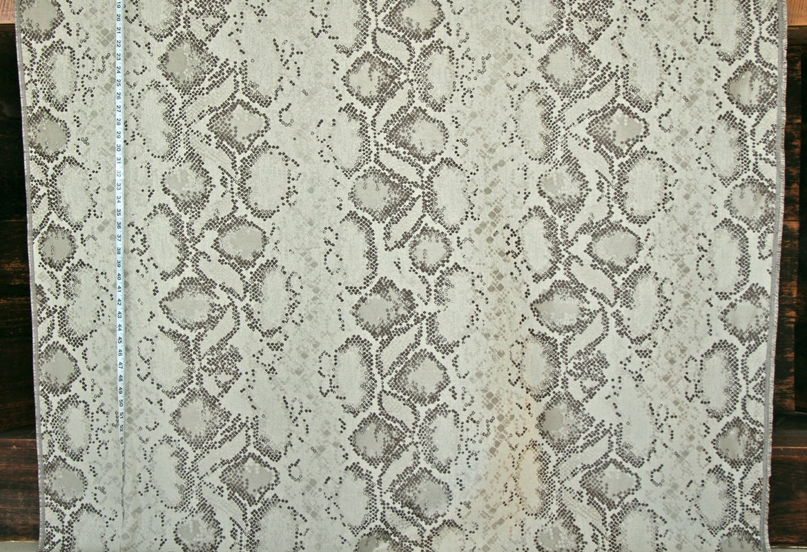 A close-up of a snakeskin-patterned fabric or wallpaper sample, showing a beige and gray reptile print with a white measuring tape vertically aligned on the left side.