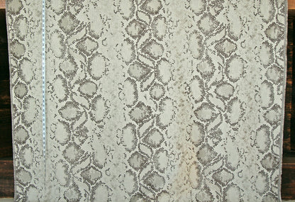 A close-up of a snakeskin-patterned fabric or wallpaper sample, showing a beige and gray reptile print with a white measuring tape vertically aligned on the left side.