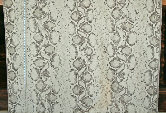 A close-up of a snakeskin-patterned fabric or wallpaper sample, showing a beige and gray reptile print with a white measuring tape vertically aligned on the left side.