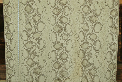 A close-up of a snakeskin-patterned fabric or wallpaper sample, showing a repeating reptile print in beige, tan, and brown tones. A white measuring tape is visible along the left edge for scale, and the material is photographed against a dark wood background.